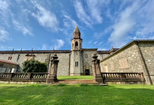 A stone building with a clock tower, surrounded by green grass and a blue sky with clouds.
