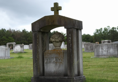 A stone mausoleum with a cross and floral decoration, surrounded by gravestones in a cemetery.