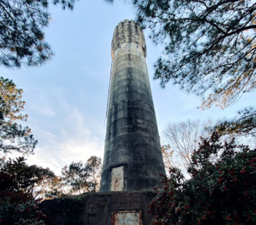 A tall, cylindrical stone tower surrounded by trees and blooming flowers against a clear sky.