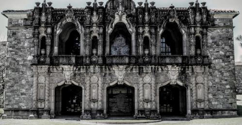 A grand, ornate stone building with intricate carvings and large arched entrances, set against a monochrome background.