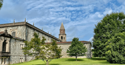 A stone building with a clock tower, surrounded by trees and a blue sky with clouds.