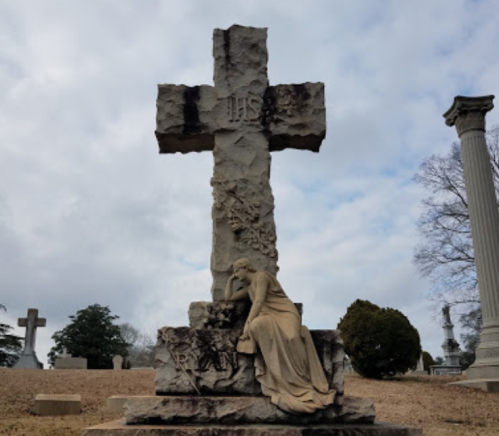 A stone cross with a seated figure in mourning, surrounded by a cemetery landscape under a cloudy sky.