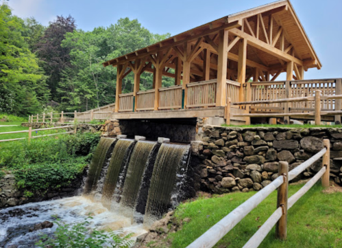 A wooden pavilion overlooks a small waterfall, surrounded by greenery and a wooden fence.