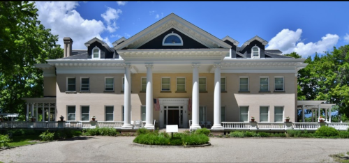 A large, elegant mansion with white columns, a dark roof, and lush greenery under a blue sky with clouds.
