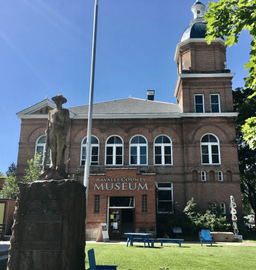 Historic Ravalli County Museum building with a statue in front, surrounded by green trees and blue benches.
