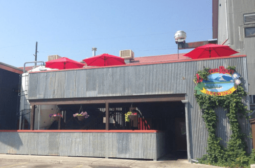 A brewery with a corrugated metal exterior, red umbrellas on the rooftop, and colorful flowers hanging from the walls.