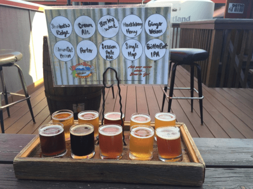 A wooden tray holds ten small beer samples, with a sign listing their names above. Outdoor setting with wooden deck.