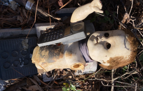 A weathered doll with a missing arm lies on the ground, holding an old mobile phone amidst leaves and debris.