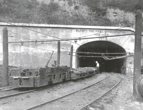 Black and white photo of a mine entrance with a cart on tracks and a worker near the tunnel, emphasizing safety.
