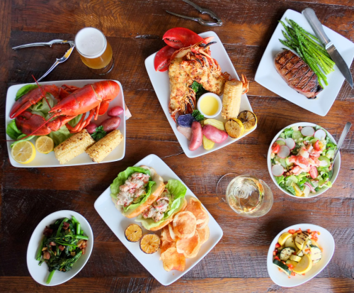 A spread of seafood, steak, salads, and sides on wooden table, with drinks and utensils.