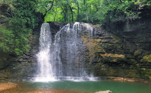 A serene waterfall cascading over rocky cliffs into a tranquil pool, surrounded by lush green trees.