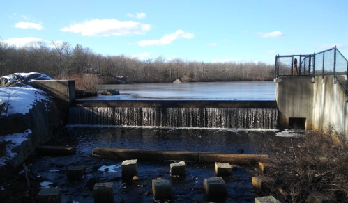 A serene view of a dam with water cascading over it, surrounded by trees and a clear blue sky.