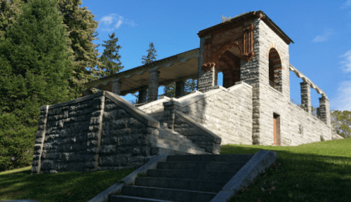 A stone structure with arches and columns, set on a grassy hill under a blue sky with scattered clouds.