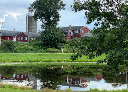 A group of people walks along a path by a pond, with red barns and a silo reflected in the water.