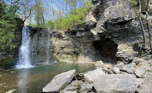 A serene waterfall cascades into a calm pool, surrounded by rocky cliffs and lush greenery under a clear blue sky.