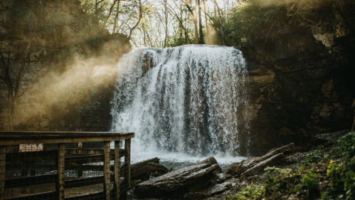 A serene waterfall cascades over rocks, surrounded by trees and soft sunlight filtering through the mist.