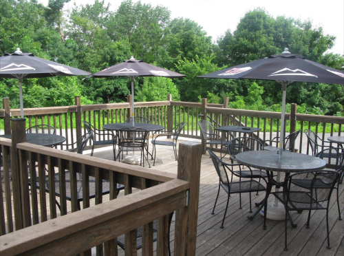 Outdoor patio with tables and umbrellas surrounded by greenery. Wooden deck and railings create a cozy atmosphere.