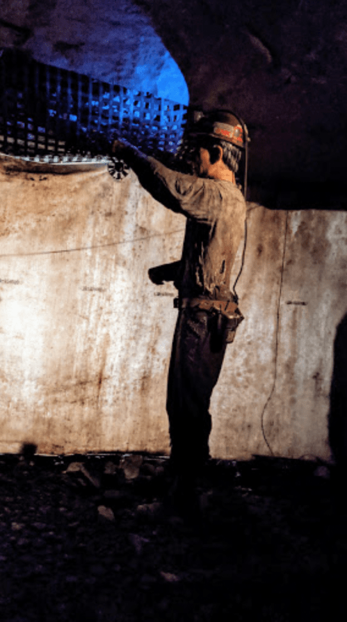 A miner in a helmet works in a dimly lit underground tunnel, using a tool near a metal grate.