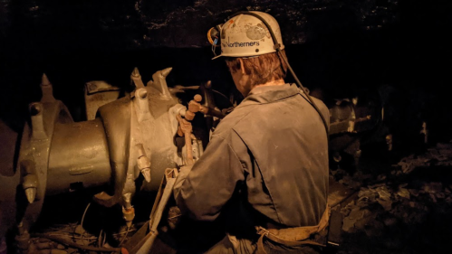 A miner in a helmet works on machinery in a dimly lit underground setting.