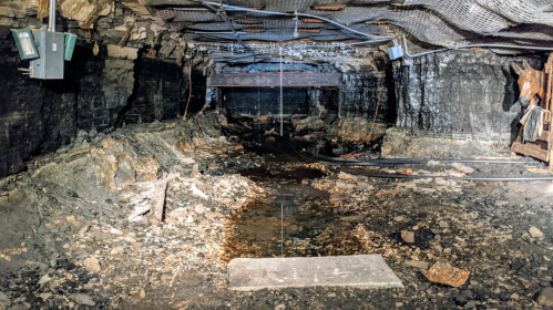 A dimly lit underground tunnel with rocky walls, water on the ground, and exposed electrical equipment.