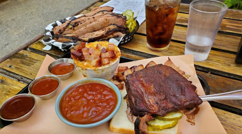 A plate of barbecue with brisket, pickles, baked beans, mac and cheese topped with bacon, and drinks on a wooden table.
