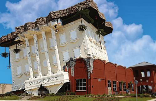 A whimsical building with an upside-down design, featuring a cracked facade and tropical plants against a blue sky.