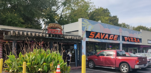 Exterior of Savage Pizza restaurant with colorful signage and a red pickup truck parked nearby. Lush greenery in front.