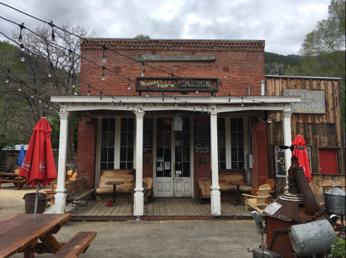 A rustic saloon with a wooden porch, red brick facade, and outdoor seating, surrounded by trees and string lights.