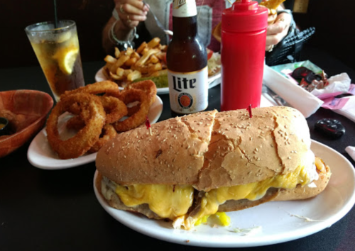 A hearty sandwich with melted cheese, onion rings, fries, a beer, and condiments on a table.