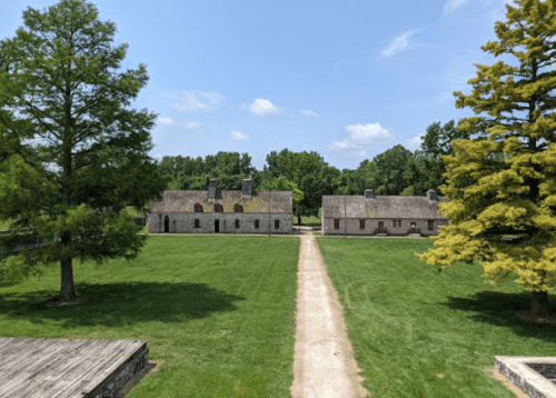 A historic site featuring stone buildings and a grassy pathway, surrounded by trees under a clear blue sky.