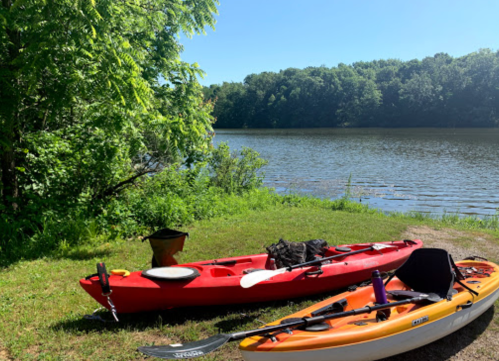 Two kayaks, one red and one orange, are parked on the grassy shore by a calm river surrounded by trees.