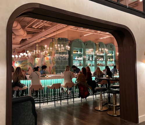 A vibrant bar scene with patrons seated at a glowing counter, colorful decor, and shelves filled with bottles in the background.