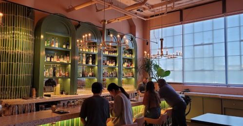 A stylish bar with green shelves filled with bottles, featuring patrons seated at the counter and warm lighting.