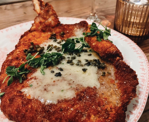 A crispy, golden-brown breaded dish topped with melted cheese, capers, and fresh herbs on a decorative plate.