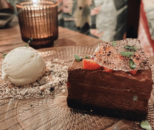 A slice of chocolate cake topped with strawberries and a crispy garnish, served with a scoop of vanilla ice cream.