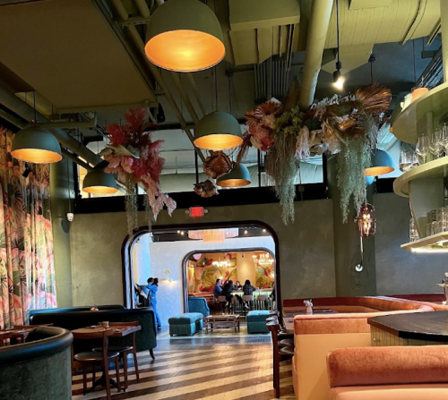 Interior of a stylish restaurant featuring colorful decor, hanging plants, and warm lighting, with diners in the background.