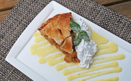 A slice of apple pie topped with whipped cream and mint leaves, drizzled with a yellow sauce on a white plate.