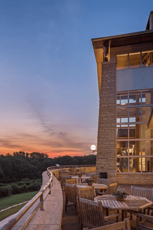 A modern stone building with a wooden deck overlooks a sunset-lit landscape, featuring outdoor dining furniture.