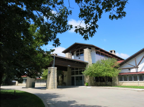 A modern building with a stone facade and large windows, surrounded by trees and a clear blue sky.