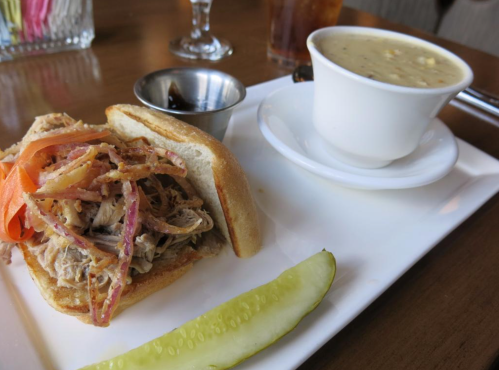 A plate featuring a pulled chicken sandwich with pickled vegetables, a cup of soup, and a pickle slice on the side.