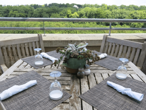 A wooden dining table set with glasses and napkins, surrounded by greenery and a scenic view.