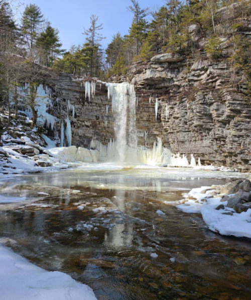 A frozen waterfall cascades into a partially frozen pool, surrounded by rocky cliffs and snow-covered trees.