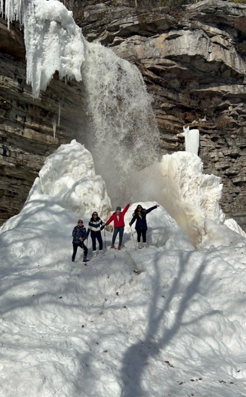 Four people stand in front of a frozen waterfall, surrounded by snow and ice, with sunlight casting shadows.