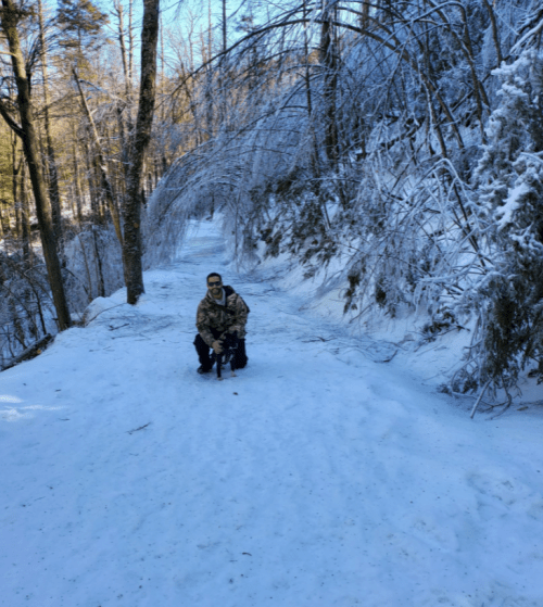 A person in winter clothing sits on a snowy path surrounded by trees, with a serene winter landscape in the background.