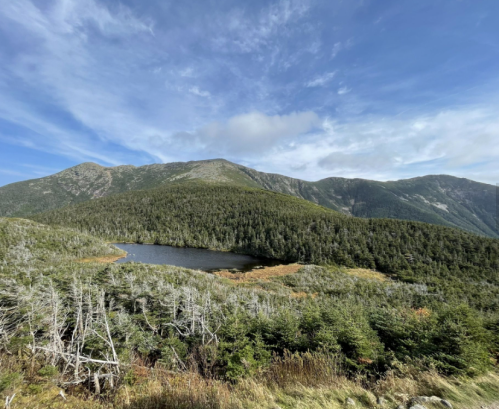 A scenic view of a mountain landscape with a lake surrounded by trees under a blue sky with scattered clouds.