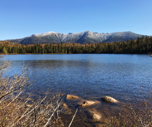 A serene lake reflects snow-capped mountains, surrounded by autumn foliage and rocky shores under a clear blue sky.