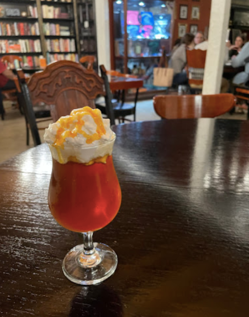 A glass of iced tea topped with whipped cream and caramel sauce, set on a wooden table in a cozy café.