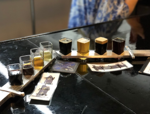 A wooden flight board with various small glasses of different colored liquids and tasting notes on a dark table.