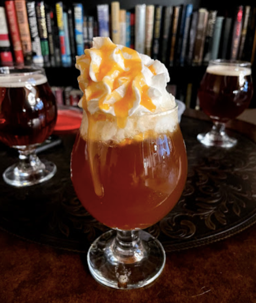 A glass of caramel-drizzled dessert drink topped with whipped cream, set against a backdrop of books and other drinks.