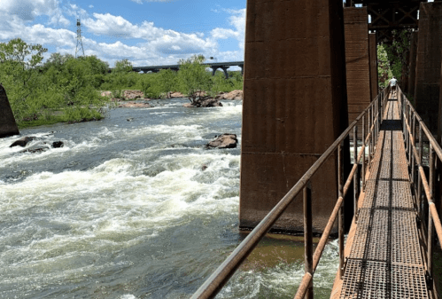 A metal walkway beside a rushing river, with green trees and a bridge in the background under a blue sky.